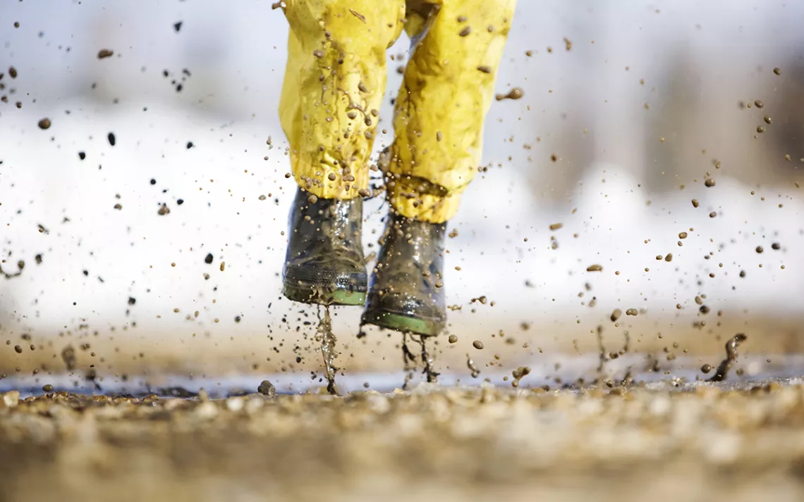 Child photographed from the waist down jumping into a puddle with yellow rain boots