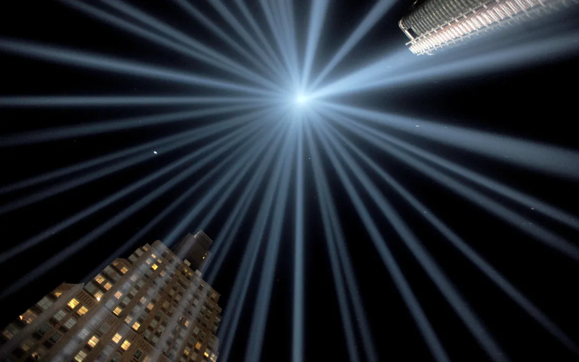 Skyscrapers against a night sky as seen from below, with spotlights on all sides meeting in the center