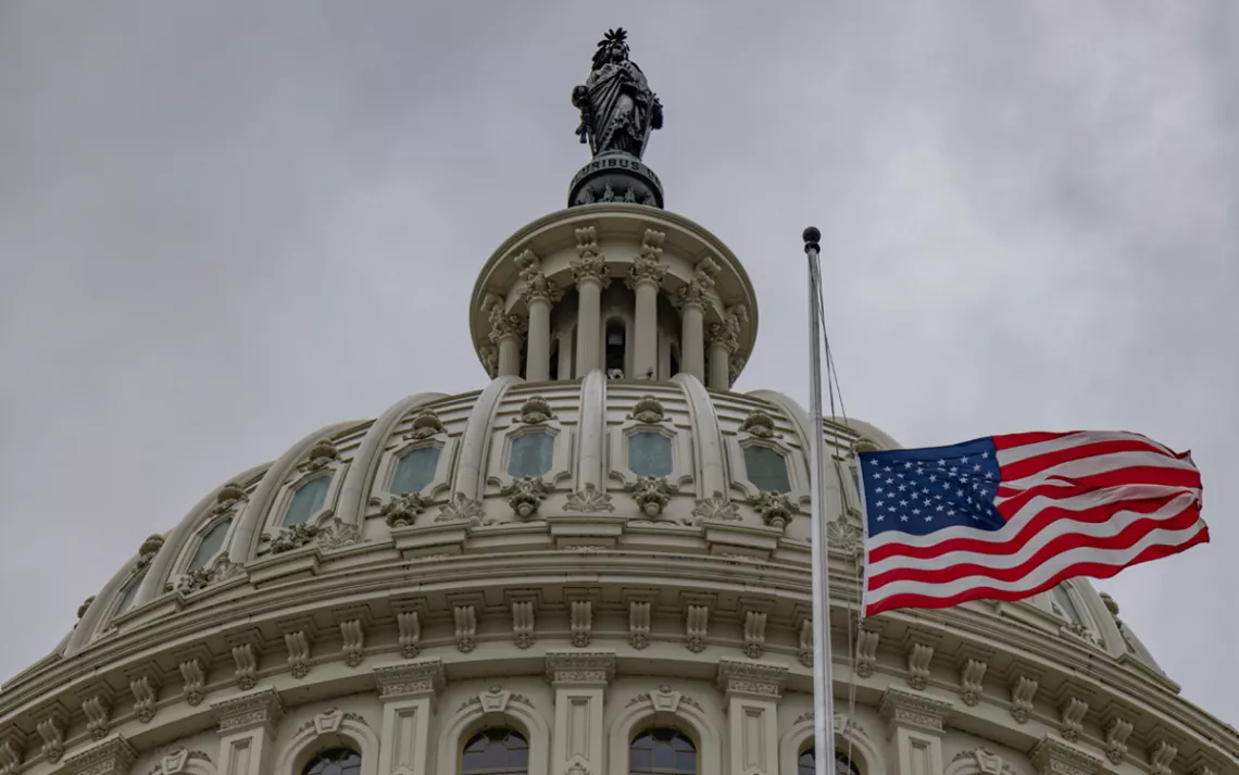 Top of the US Capitol building, with an American flag flying out front and a cloudy sky overhead.