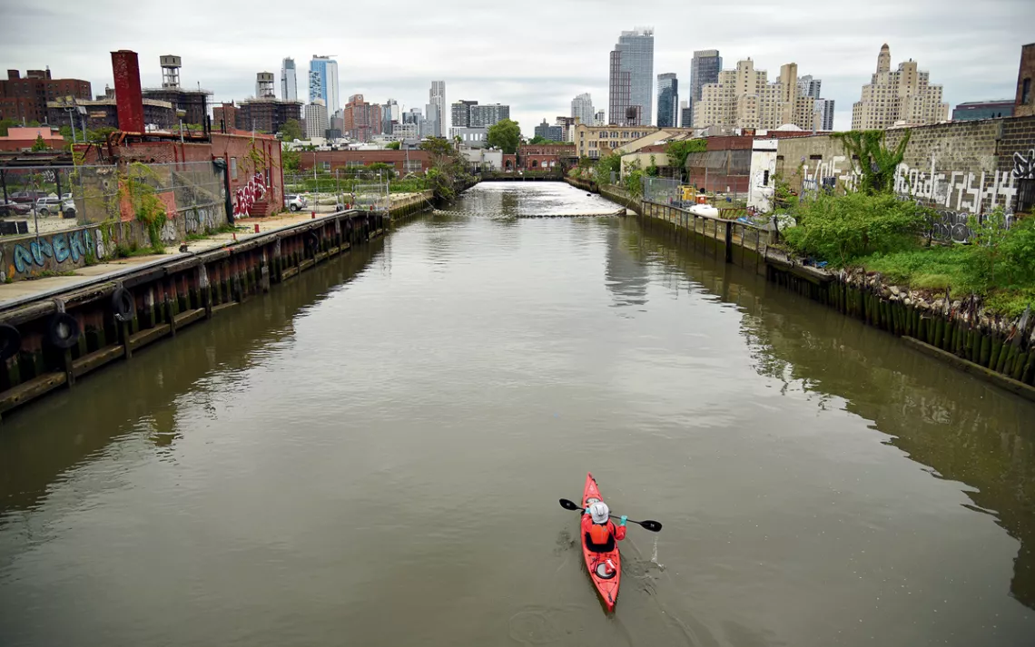 A red kayak floats down a brown, murky canal with industrial buildings along the sides and tall city buildings in the distance.
