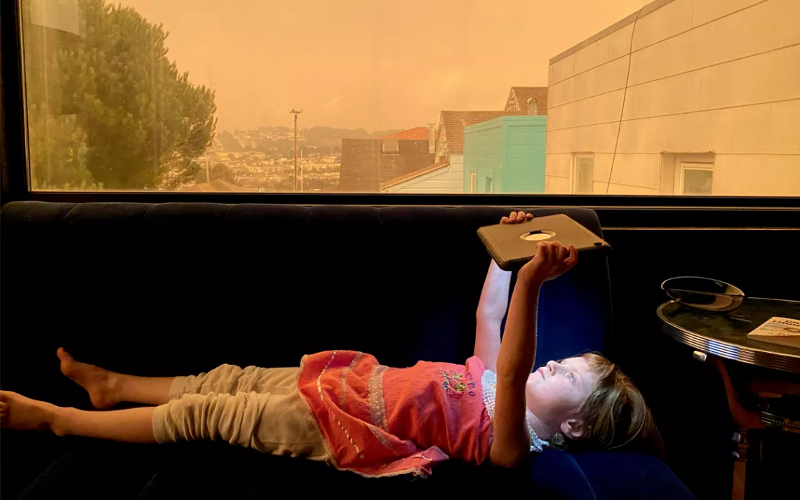 A young girl lies inside on a bench by a window holding an iPad over her head. Above her, out the window, the sky is orange from wildfire smoke.