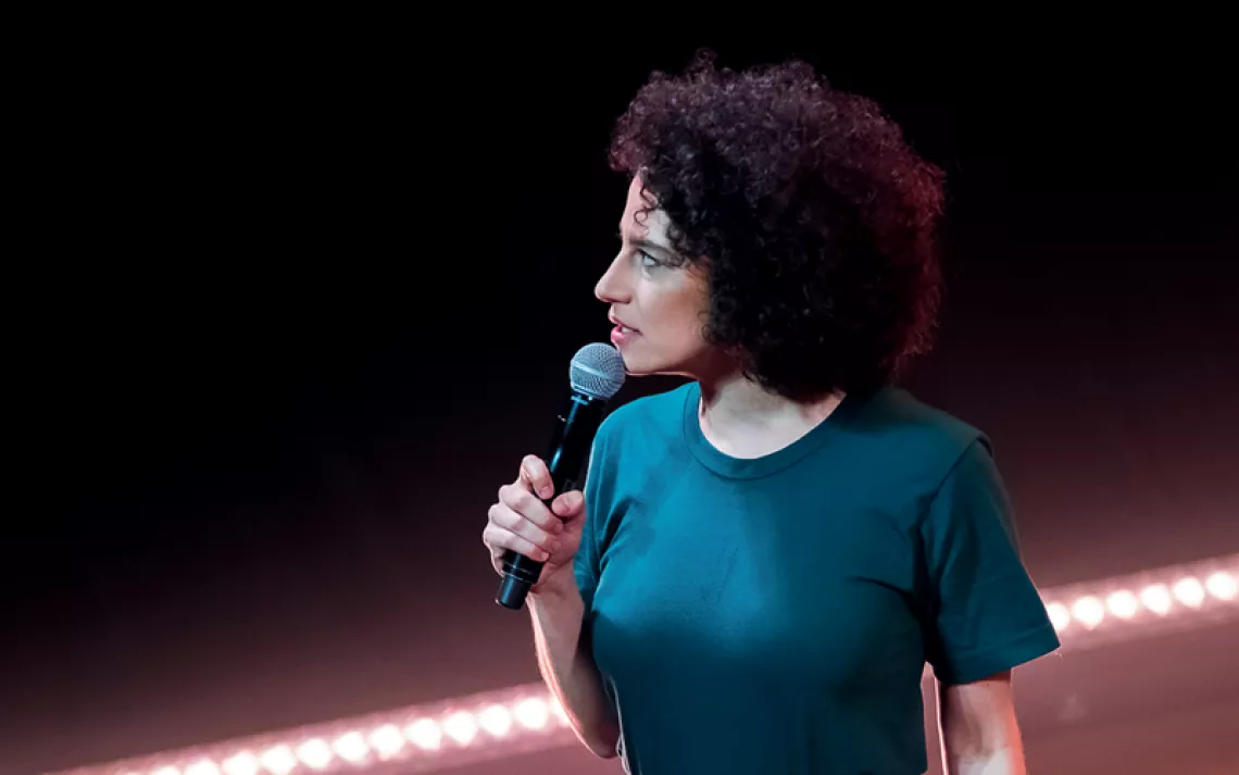 Ilana Glazer is on stage, holding a mic and looking to her right. She's wearing cut-off jean shorts and a teal T-shirt.