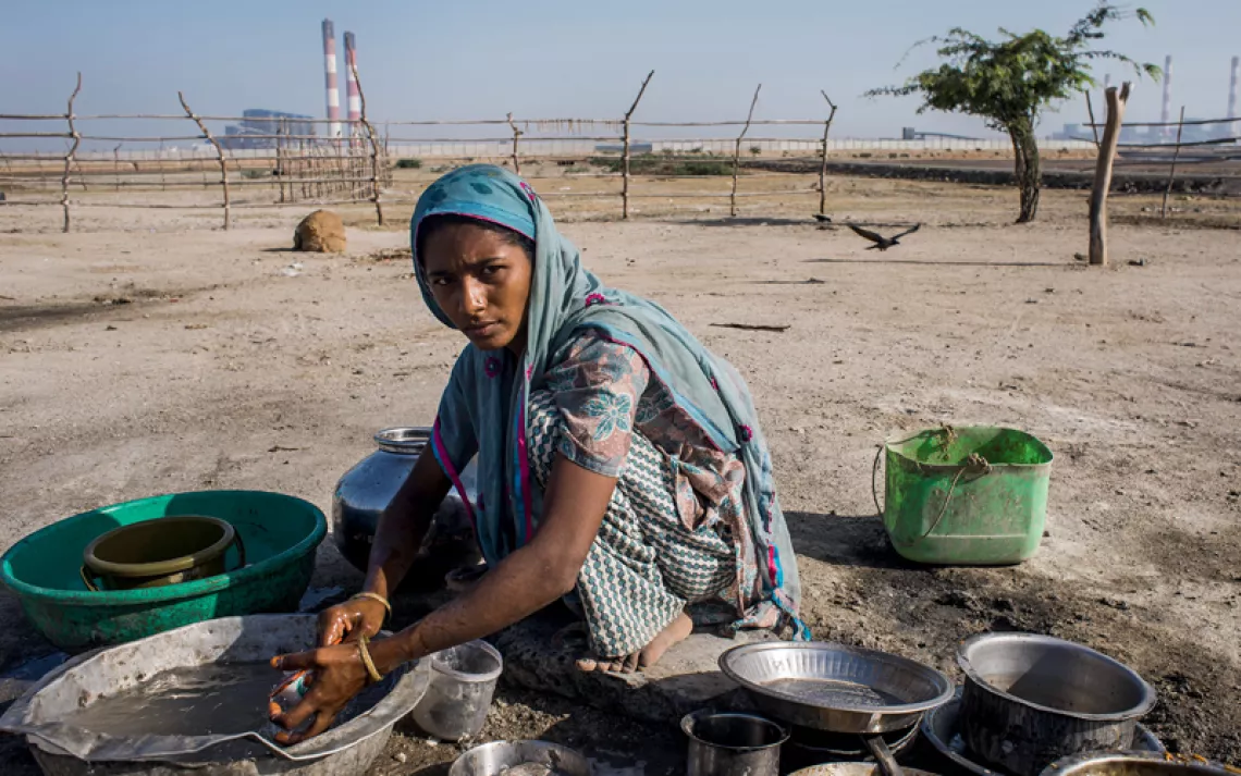 A woman wearing a scarf and Indian clothing washes dishes in a bowl outside. The Tata Mundra power pl​ant is in the background.