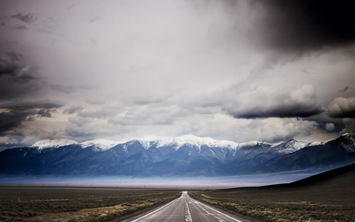 Looking down Nevada's Route 50, with no cars, snowy mountains in the background, and a cloudy sky