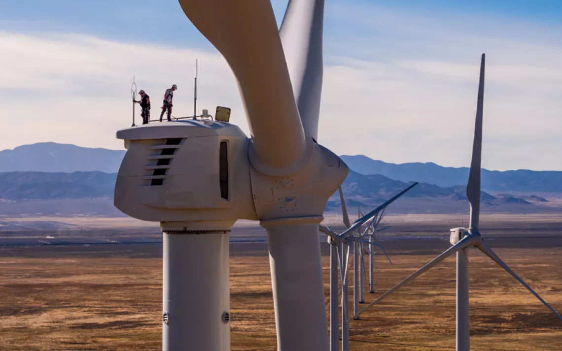 Two men stand atop a wind turbine at Utah's Milford Wind Corridor wind farm. To the right are several other turbines. They're sitting on a large plain with small hills in the background.