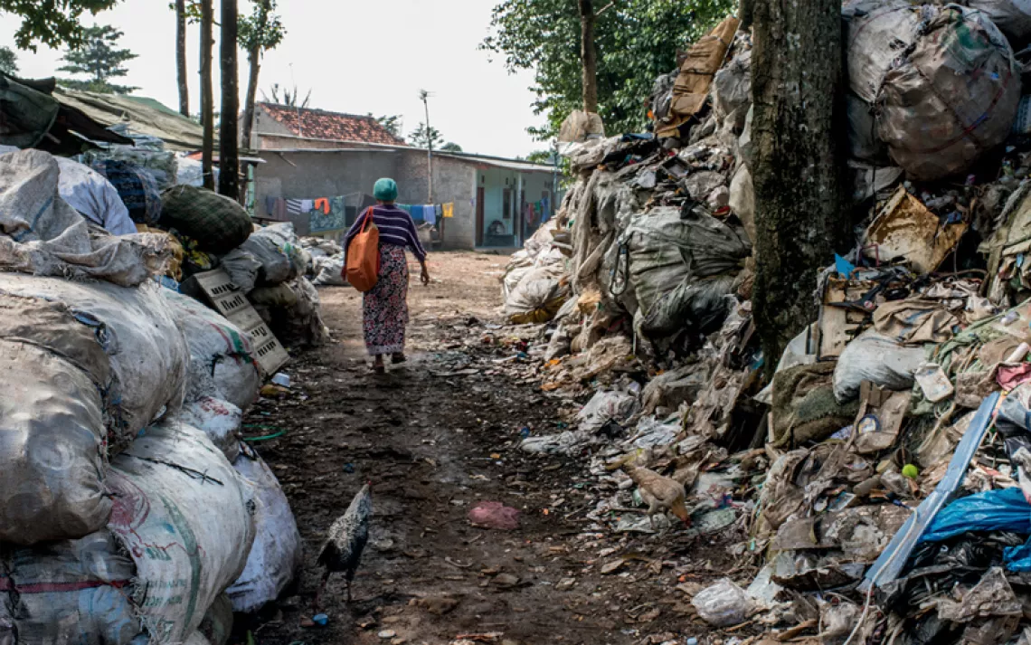 The back of midwife Mak Muji walking down a path toward a building in between two large piles of garbage and trees. In a bag, she's carrying the analog scale she uses to weigh babies.