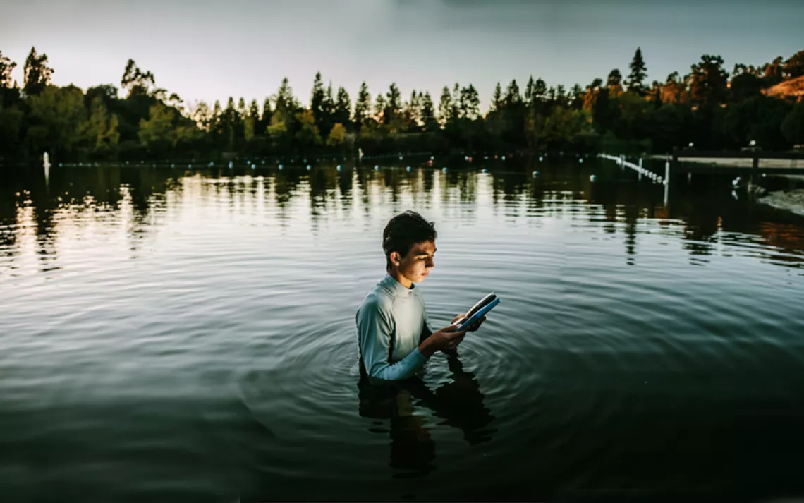 A young man stands in Lake Temescal, Oakland, at dusk holding an iPad. There's a soft glow on his face.