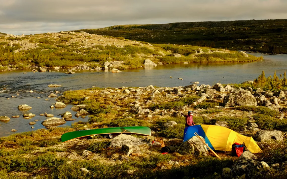 A woman stands by a yellow and blue tent and a green canoe in front of the Inukjuak River in Quebec's Tursujuq National Park.