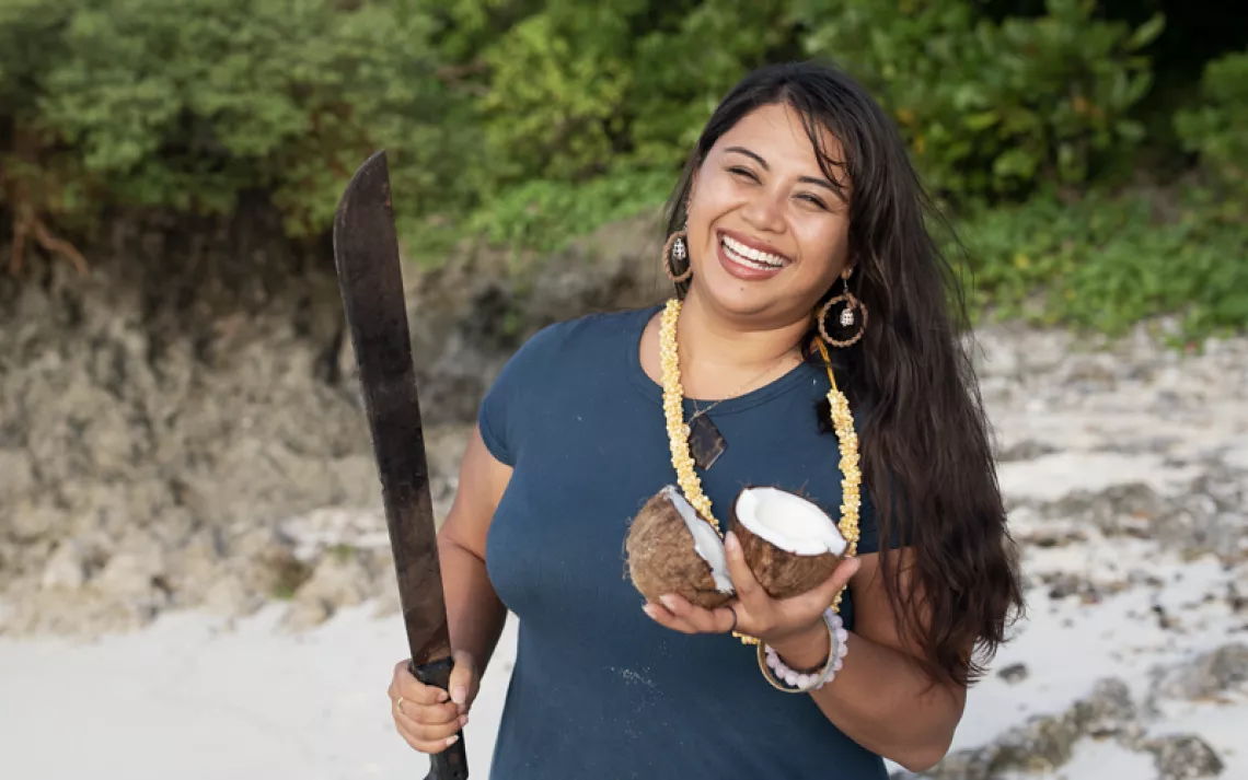Moñeka De Oro smiles and stands on a beach holding a cut coconut and a machete.