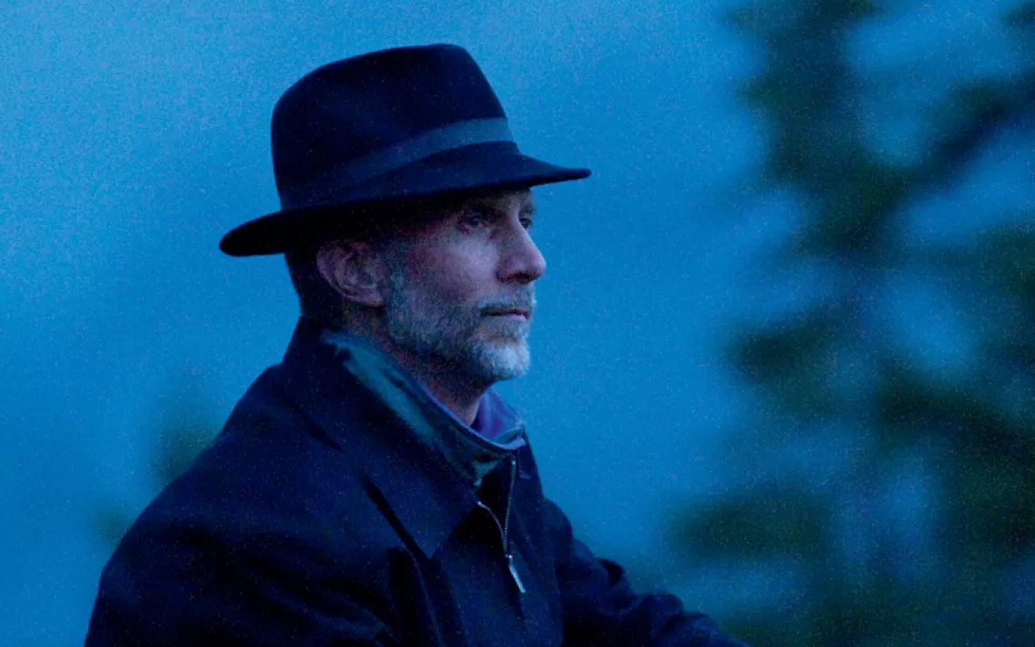A dark photo of John Luther Adams in a fedora looking off in the distance.