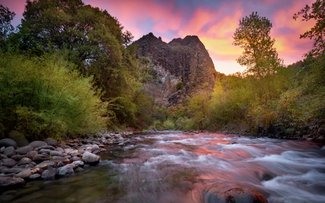 A river flows through the Ishi Wilderness in California. The sky is multicolored, and the river is lined with rocks and trees.