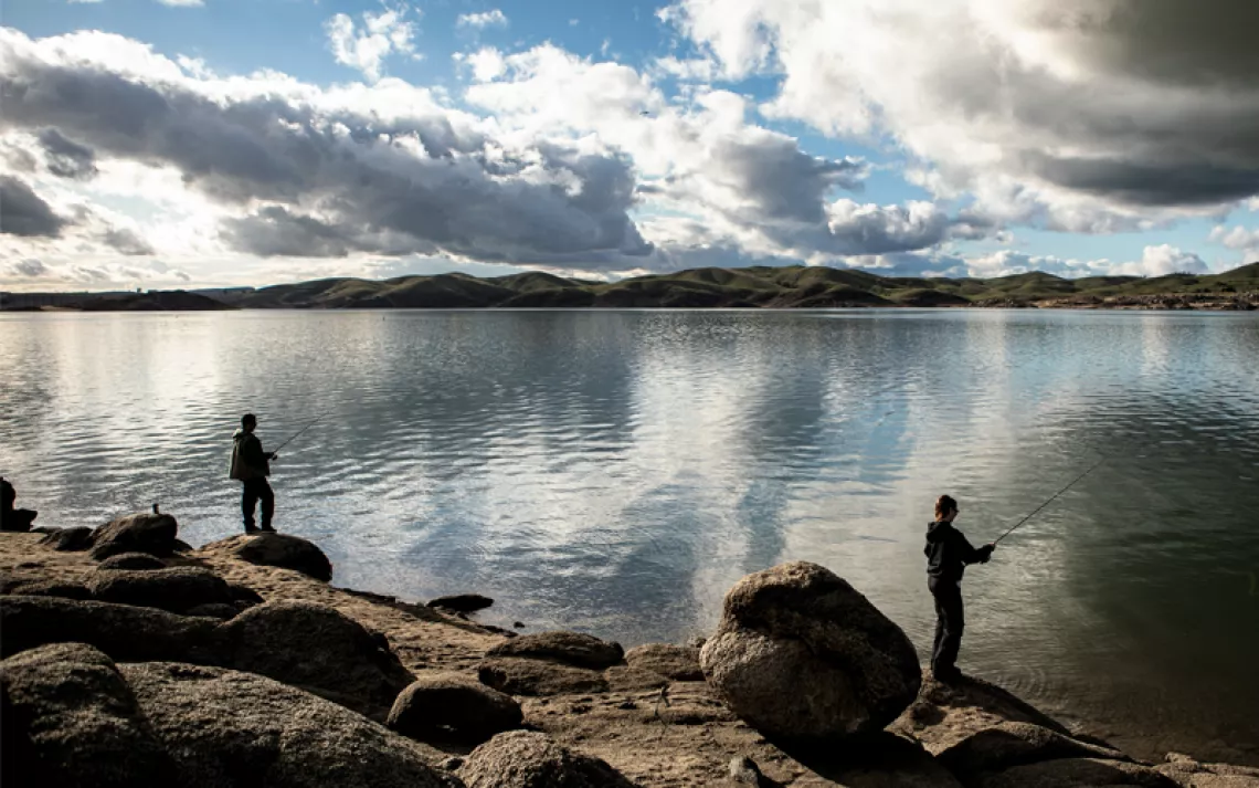 Two people are fishing from the shore of Millerton Lake. The water is glassy, and blue sky pokes through clouds above.