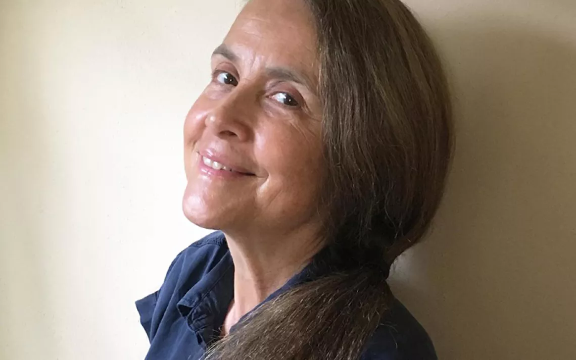Naomi Shihab Nye looks to the left over her shoulder and smiles at the camera. Nye has long, dark hair and is wearing a dark-blue button-up shirt.