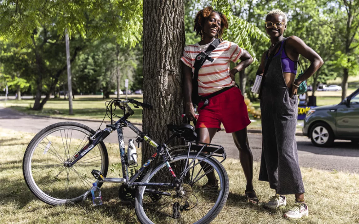 Two women wearing summer clothes pose in front of a tree in a park, smiling. A bike is leaned up against the tree.
