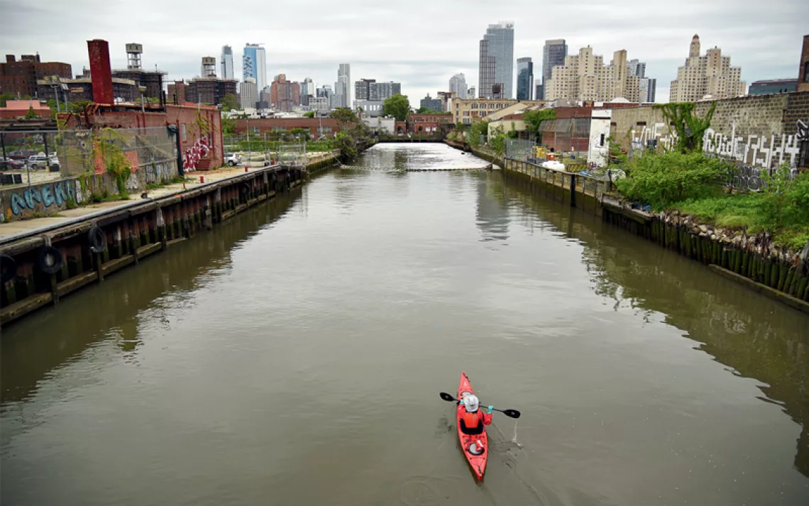 A red kayak floats down a brown, murky canal with industrial buildings along the sides and tall city buildings in the distance.