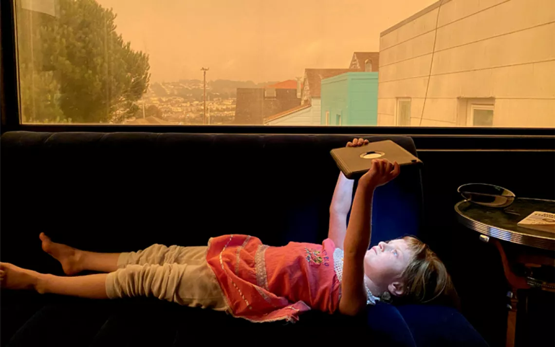 A young girl lies inside on a bench by a window holding an iPad over her head. Above her, out the window, the sky is orange from wildfire smoke.