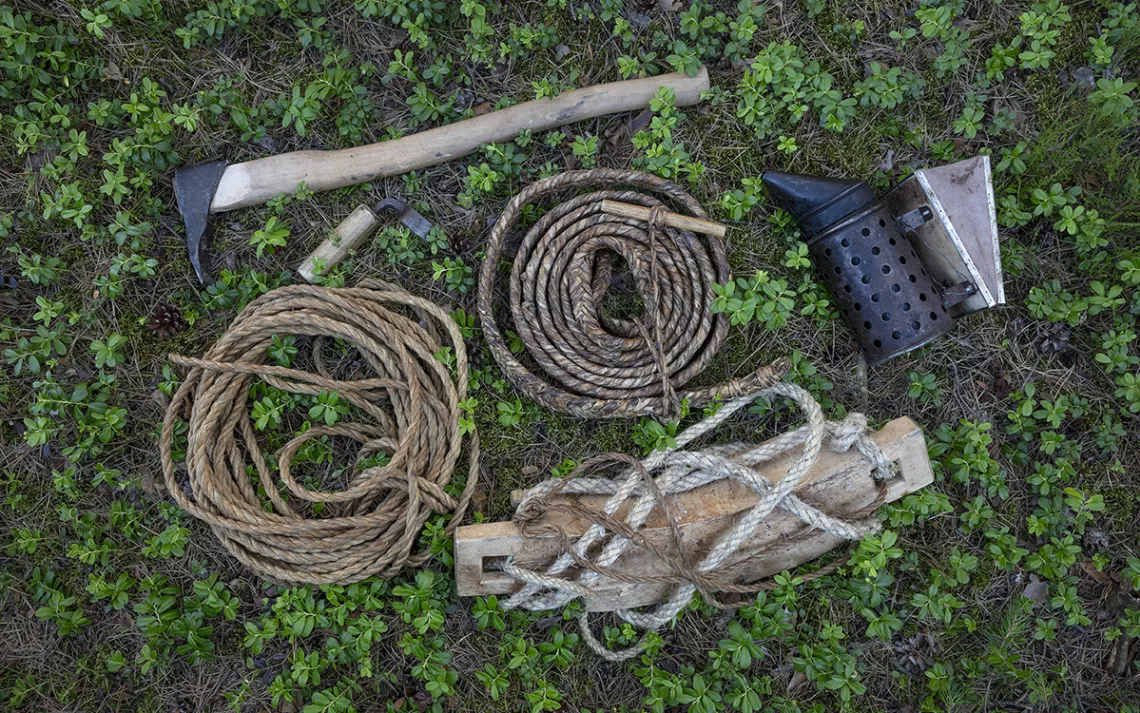 A collection of tools lying in the green grass: among them a small pickaxe, several handmade ropes, and a smoker. 