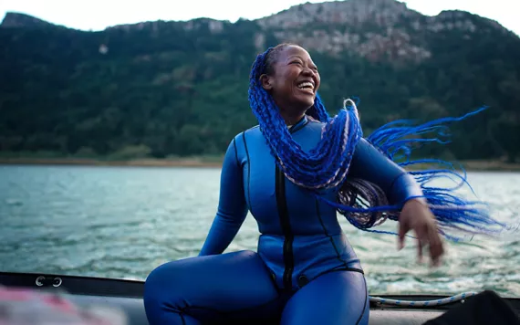 A still from the film The Black Mermaid, featuring a Black woman in a purple wetsuit with long purple braids out on the water, living life to the fullest. 