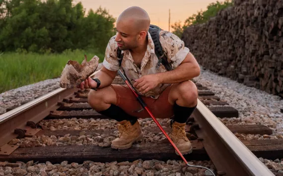 Alexi Grousis squats down in the middle of some train tracks. He has a metal hook tool and is holding up what looks like an old turtle shell.