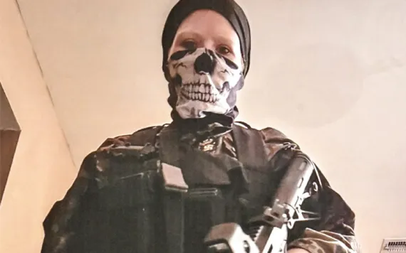 Sarah Beth Clendaniel wears camouflage clothing, a black head wrap, and a skeleton face mask. She's looking down at the camera and holding an assault rifle.