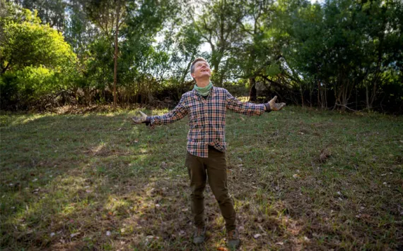 Ole Schell stands in a grassy field with a plaid shirt and his arms spread, smiling and looking at the sky.