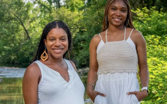 Diamon Clark and Kristen Walker stand by a tree-lined creek wearing long white dresses.