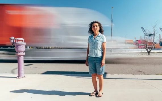 Alicia Rivera is wearing a light blue shirt, jean shorts, and flip flops and standing in front of traffic by the port.