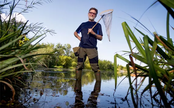 Dick Jordan smiles at the camera and holds a net while wearing tall wading boots and standing in a grass-lined body of water.