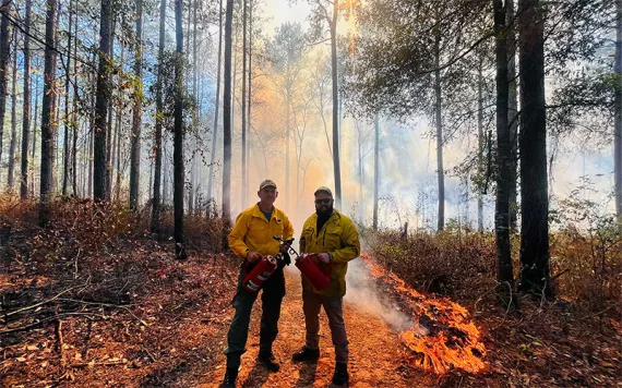 Two men dressed in yellow PPE and holding fuel canisters set a prescribed burn to a forest in Southwest Georgia.