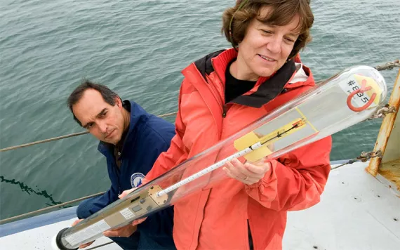 Amy Bower and an engineer hold a subsurface buoy, while standing on a ship deck, the ocean behind them.
