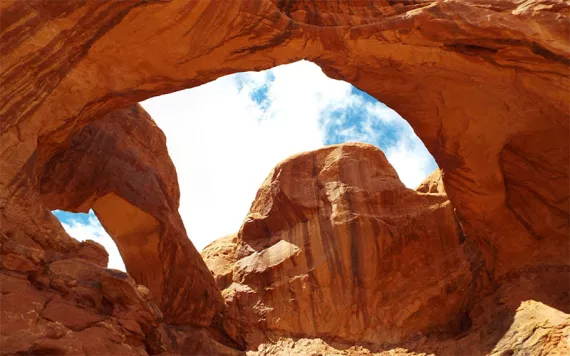 Two connected red-orange sandstone arches in front of a blue and white cloudy sk