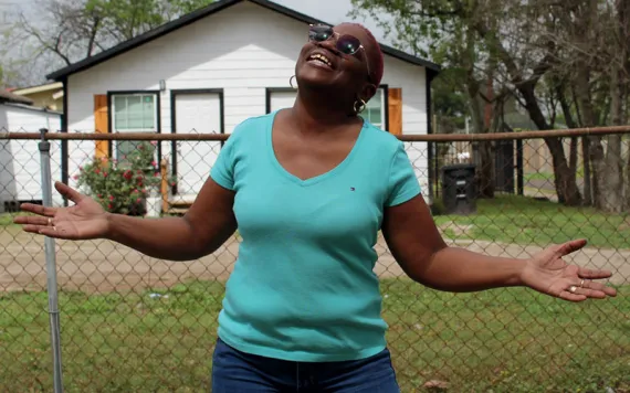 Sandra Edwards wears a turquoise shirt and sunglasses and holds her hands up to the sky and smiles. Behind her is a chain-link fence and a small white house.