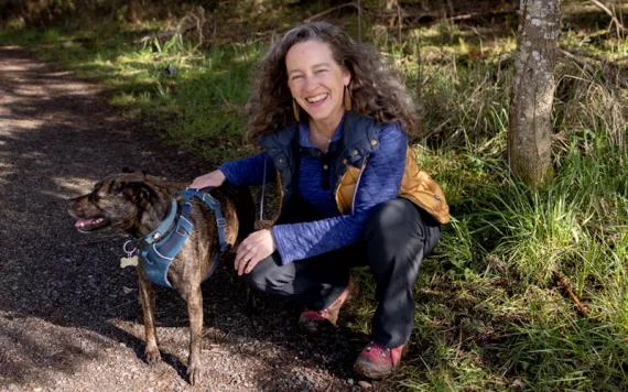  Julia Olson wears hiking clothes and smiles while kneeling down with her brown dog on a trail.