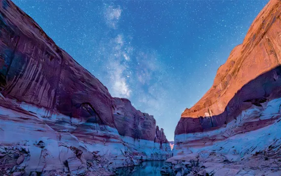 Early evening photo of a canyon in a low Lake Powell, with a white layer at the bottom and a redrock layer up top. A small boat is beached on the shore.