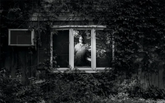  Black-and-white photo of Sean Guthrie looking at the camera through a window inside his house. The house has vines covering it.