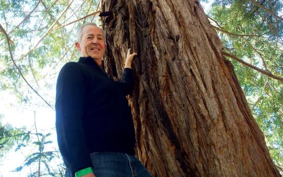 Rick Lanman stands with his left hand on a redwood tree trunk as he looks down at the camera.
