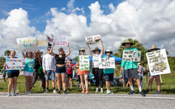 A group of protestors stand outside in sunny Florida, holding signs