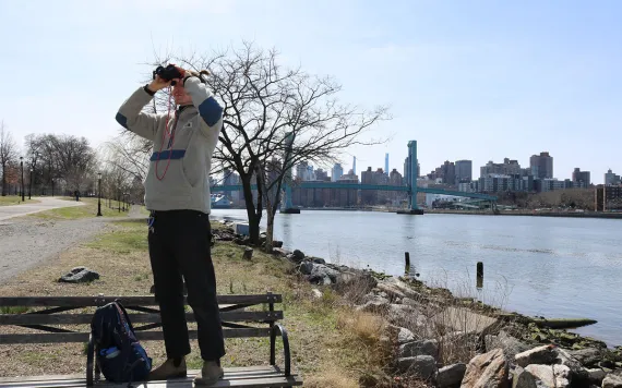 A man stands on a bench near a river, pointing binoculars toward the sky. 