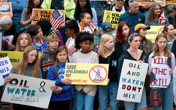 Students and adults of different races stand in front of Florida’s Department of Environmental Protection holding signs including "Oil and Water Don't Mix," "Kill the Drill," and "Save Apalachicola Bay."