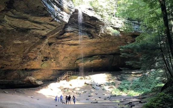 Sunlight pours through the trees onto a wide-angle view of Ash Cave with a trickle of water falling 