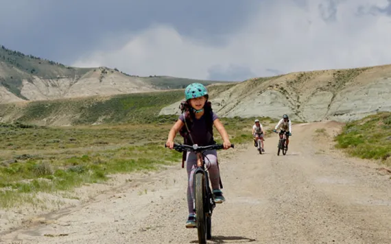 Miriam Peterson wears a turquoise helmet and rides on an unpaved road toward the camera with two other riders and the gray, rocky Oregon Buttes behind her.