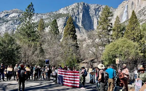 A line of protesters hold signs and an upside-down US flag in Yosemite National Park in front of a waterfall.