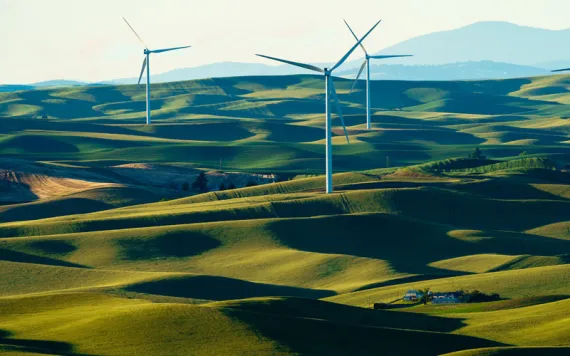 Three windmills stand to attention in a lush, rolling green field, with hills and mountains in the background