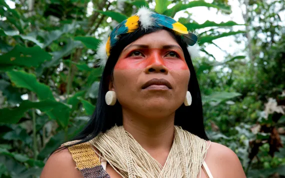 Close-up of Nemonte Nenquimo looking into the distance, wearing a feather headdress and string top. A red stripe is painted across her eyes and nose.