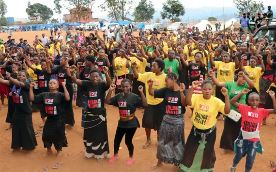 More than 100 Black women wearing different-colored "V20 1 Billion Rising" T-Shirts dance with their arms in the air on a reddish-dirt ground.