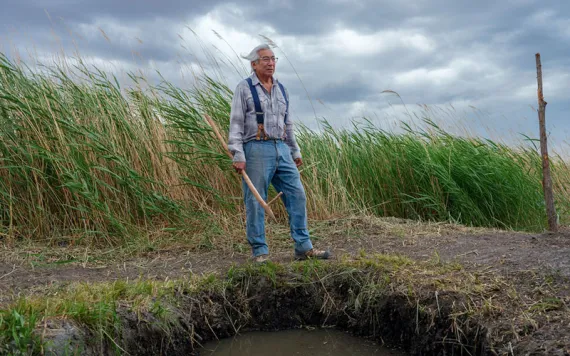 Jim Enote wears jeans and suspenders and stands atop a spring surrounded by tall grass. He's holding a long wooden instrument.