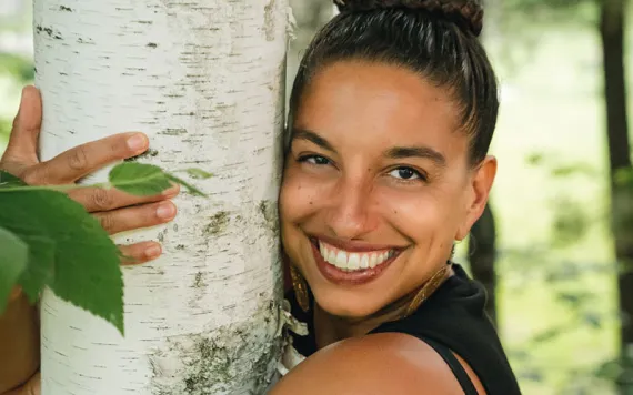 Leah Penniman wears a black top and smiles at the camera while hugging a white-barked tree.