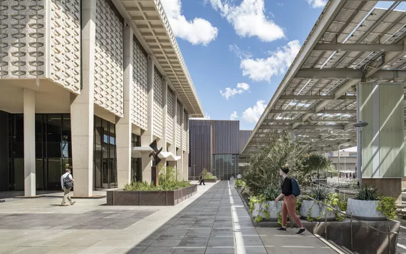 Students walk on a pathway next to a 1950/60s concrete building. To the right is a newly built outdoor area protected by panels that may be solar.