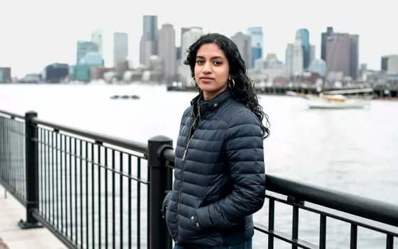 Varshini Prakash, wearing a dark-blue puffy jacket, looks into the camera while standing against a metal fence. Behind her is water and a city skyline.
