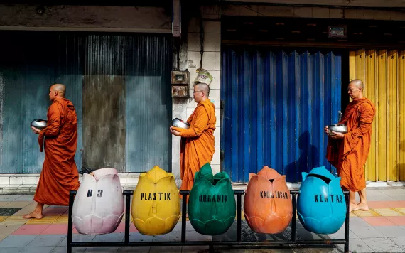 In Indonesia, three barefoot monks wearing saffron robes walk in a line, carrying silver pots for collecting alms. They're passing five bins shaped like tulips, which say (left to right) B3, Plastik, Organik, [illegible], and Kertas.